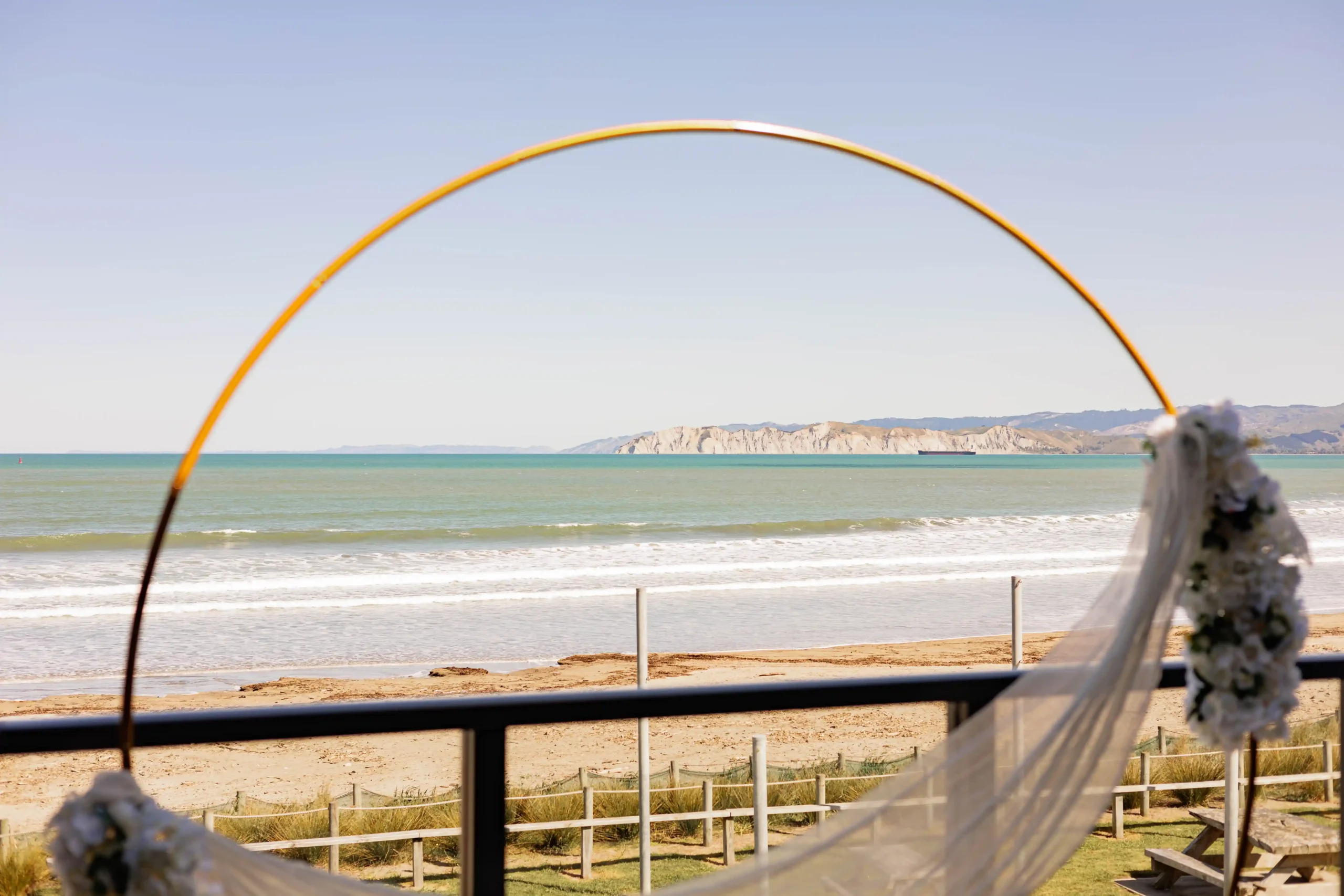 Gold floral hoop arch framing Pacific Ocean view at Gisborne beachfront wedding ceremony
